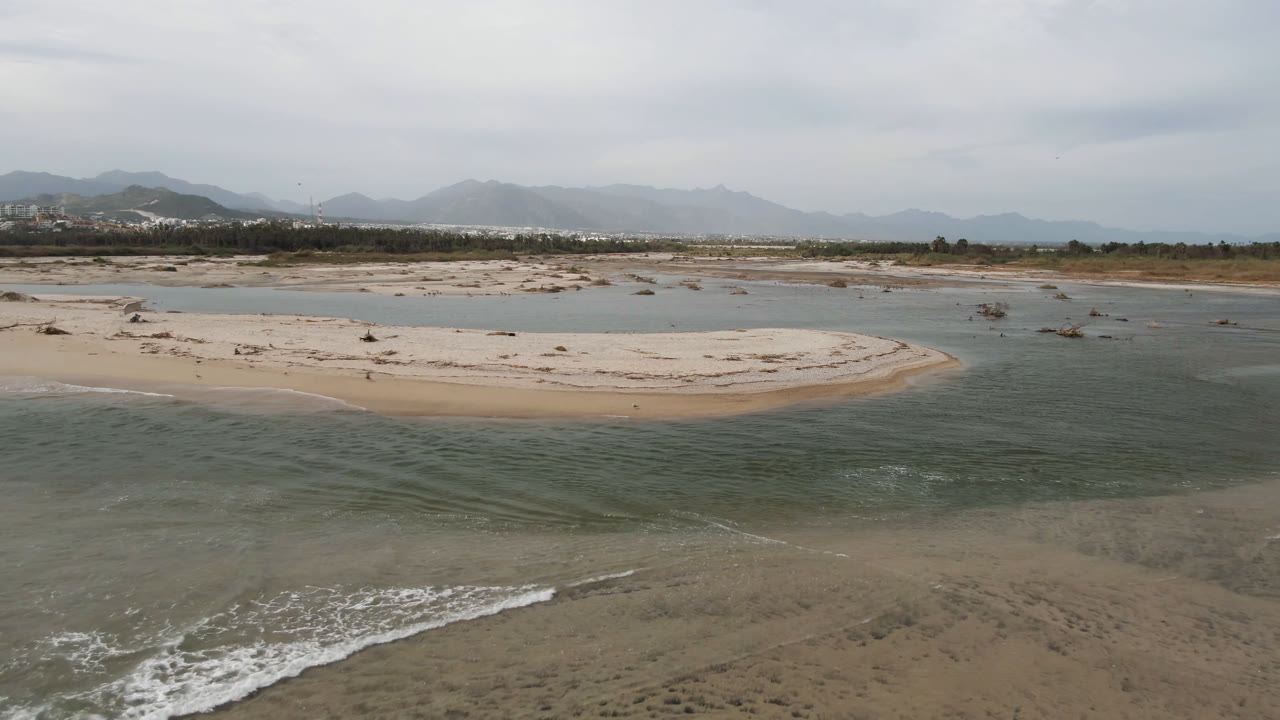 At 0:01, the drone-height view holds on green-gray ocean water, a curved sandbar, lagoon channels, distant shoreline development, mountains, cloudy sky, and no visible people.