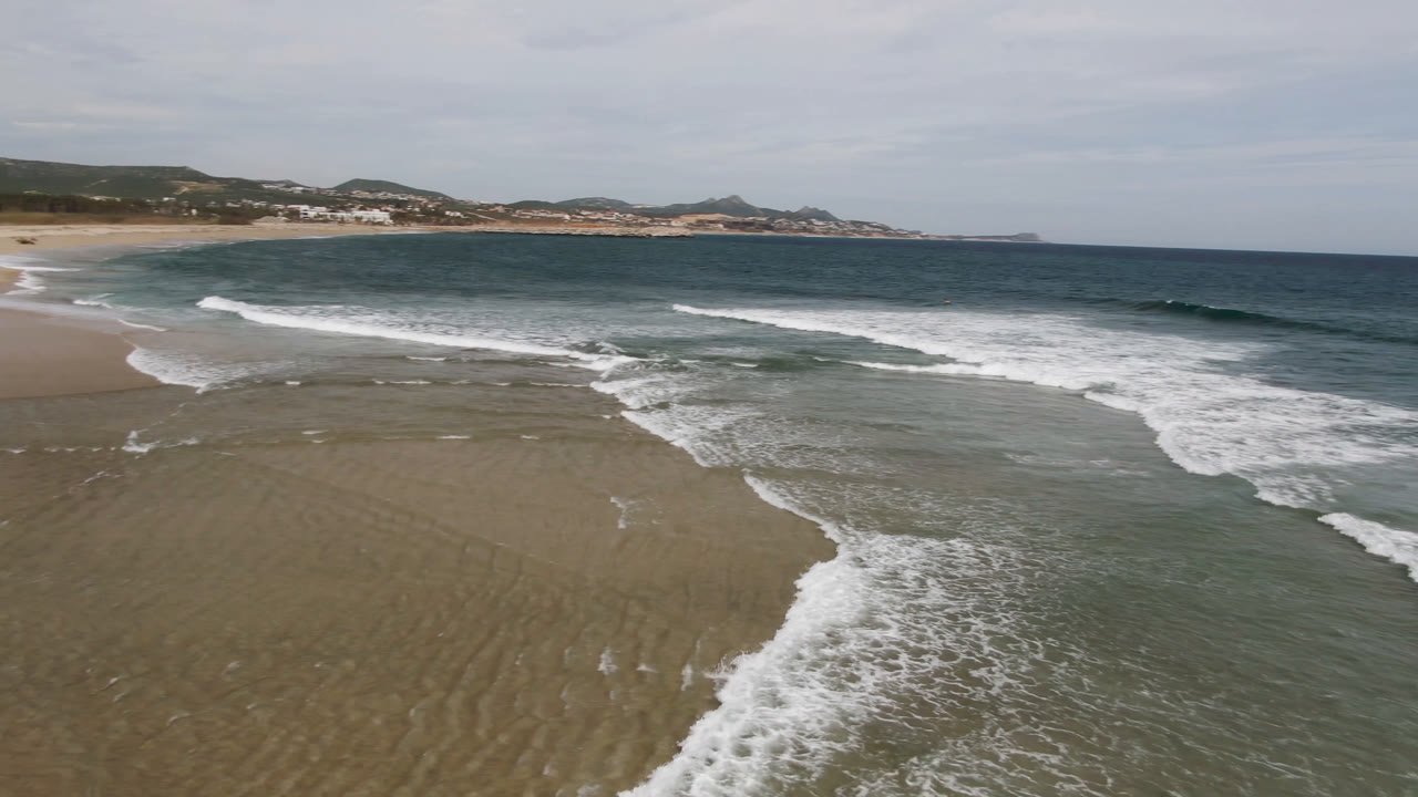 Elevated view of surf spreading over wet sand on a San Jose del Cabo beach, with a curved shoreline, distant hills, and no visible people.