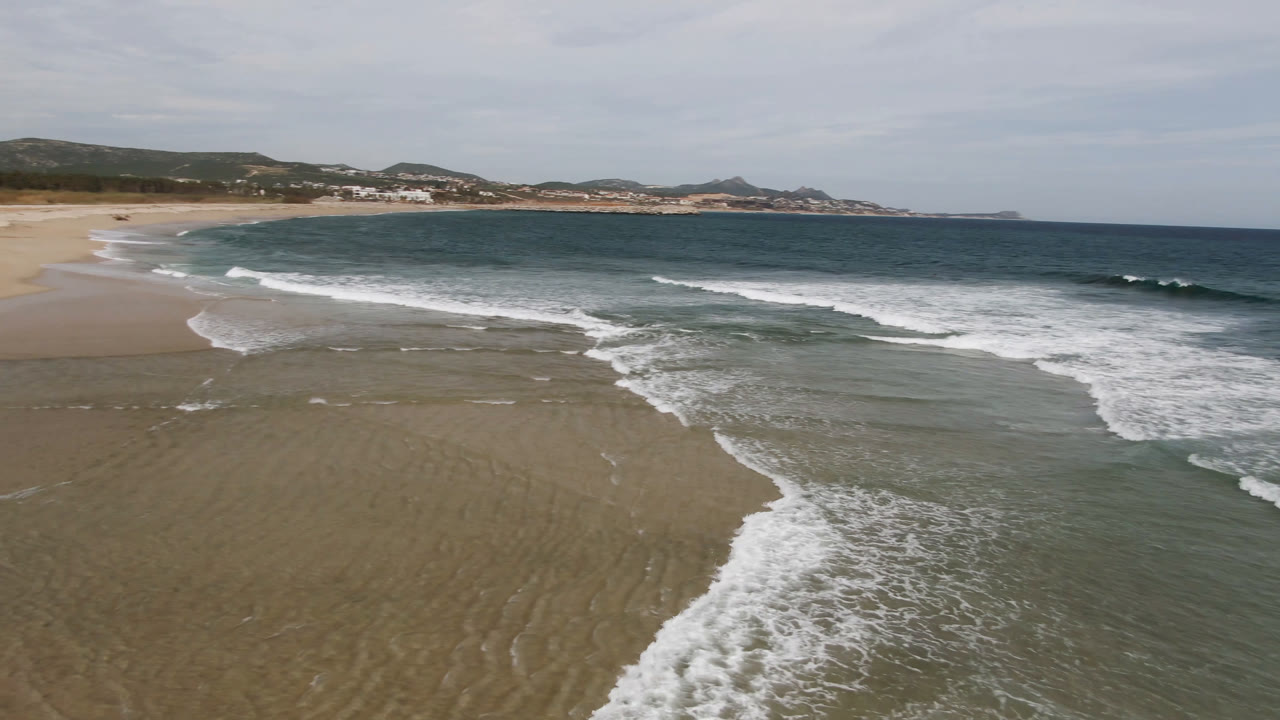 Aerial beach frame in San Jose del Cabo with a diagonal band of foam crossing shallow water, open sand to the left, distant hills, and no visible people.