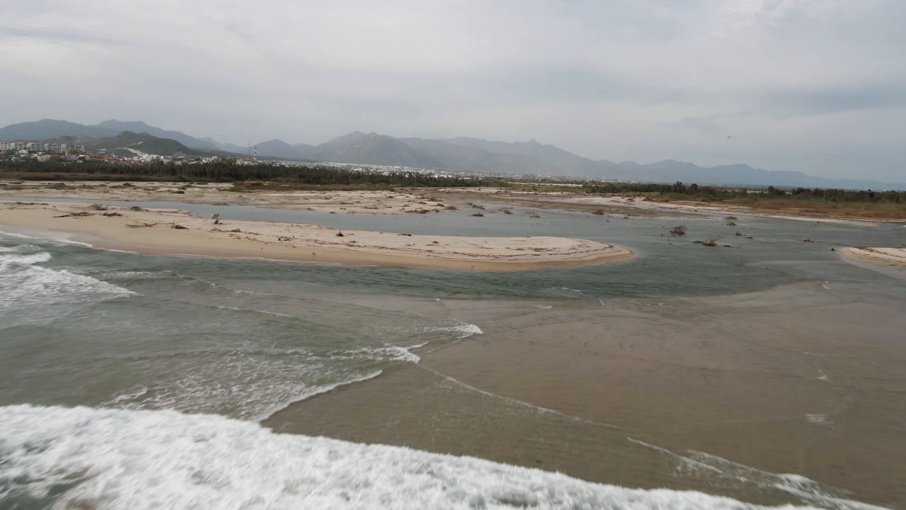 Aerial frame over the San Jose del Cabo shoreline with gray-green surf in the foreground, pale sandbars across the beach, hazy mountains beyond, and no visible people.