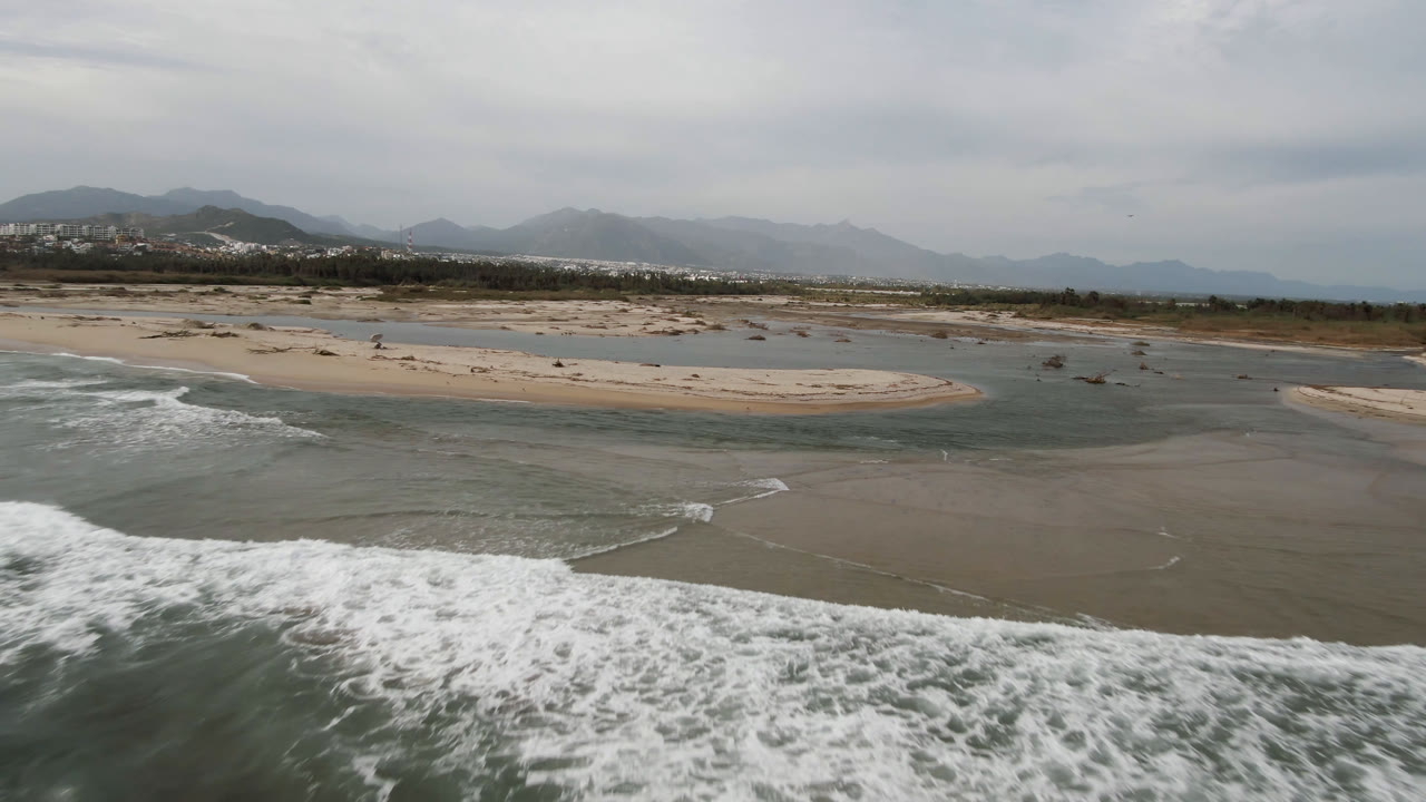 Drone frame from San Jose del Cabo with a stronger band of white foam crossing the foreground water, sandbars and beach behind it, distant town edges, mountains, and no visible people.