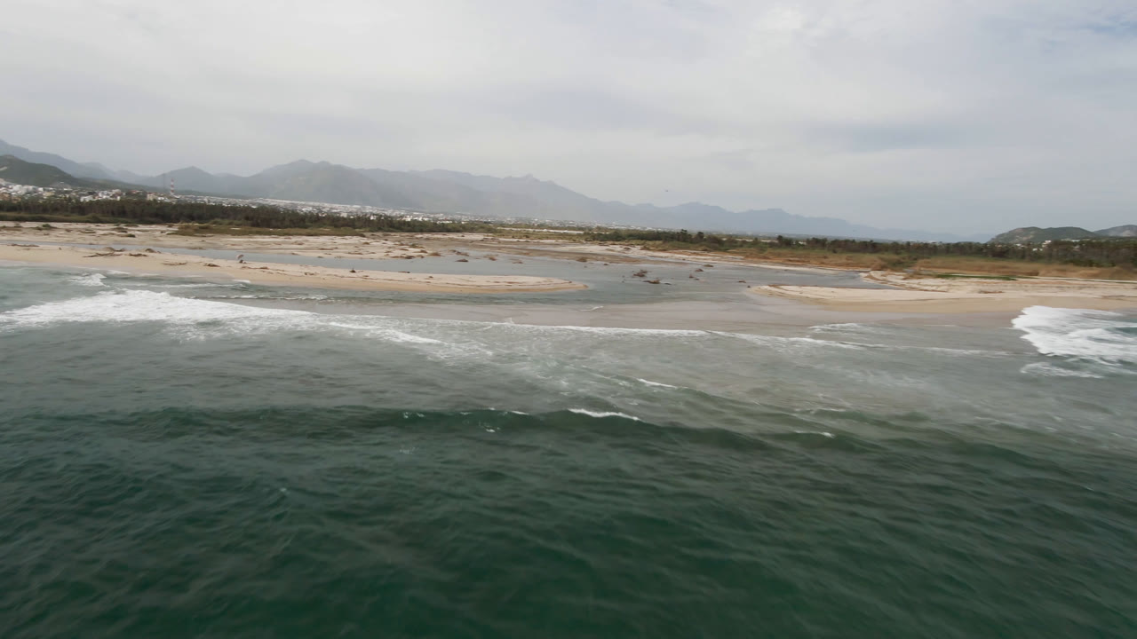 Aerial view in San Jose del Cabo, Baja California Sur, with dark green ocean in the foreground, white surf on a sandy beach, distant mountains, and no visible people.