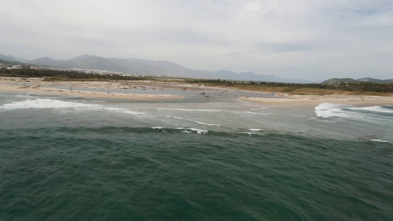 Wide aerial frame in San Jose del Cabo showing rippled ocean water, a breaking surf line, pale sand, low coastal hills, and no visible people.