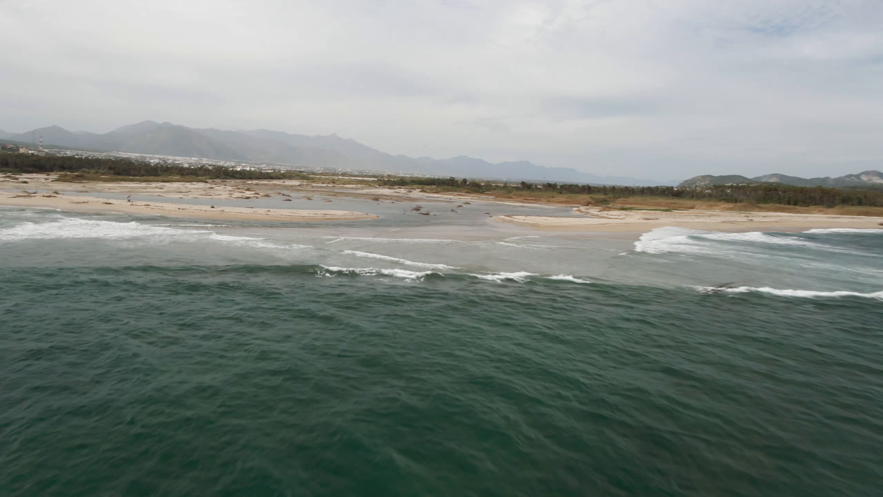 Aerial coastline with green water, white wave foam, sand, and hazy mountains.