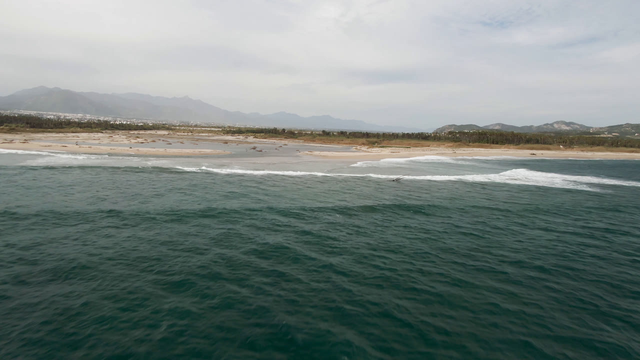 Aerial view in San Jose del Cabo, Baja California Sur, looking across dark green ocean toward white surf, sandy beach, low hills, and hazy mountains, with no visible people.