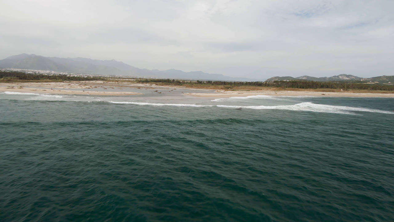 Drone-style view in San Jose del Cabo, Baja California Sur, with a broad band of ocean in front, white waves rolling toward the beach, and muted mountains under a pale sky, with no visible people.