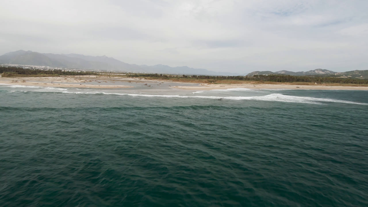 Wide aerial coastline with choppy ocean, white surf, beach, and mountains.