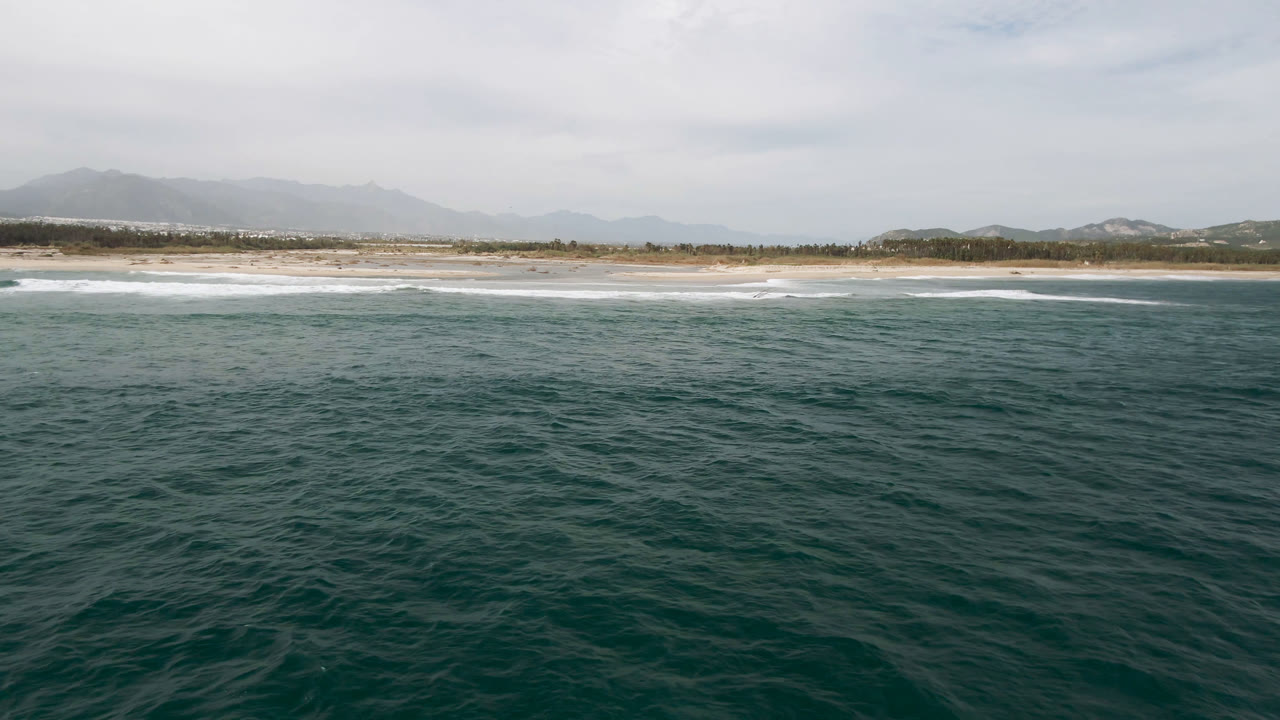 Wide aerial view of dark ocean water, surf, beach, and distant mountains.