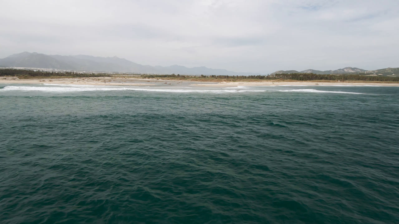 Aerial view in San Jose del Cabo with textured teal water across the foreground, a broken white surf band at the shore, low vegetation behind the sand, and no visible people.