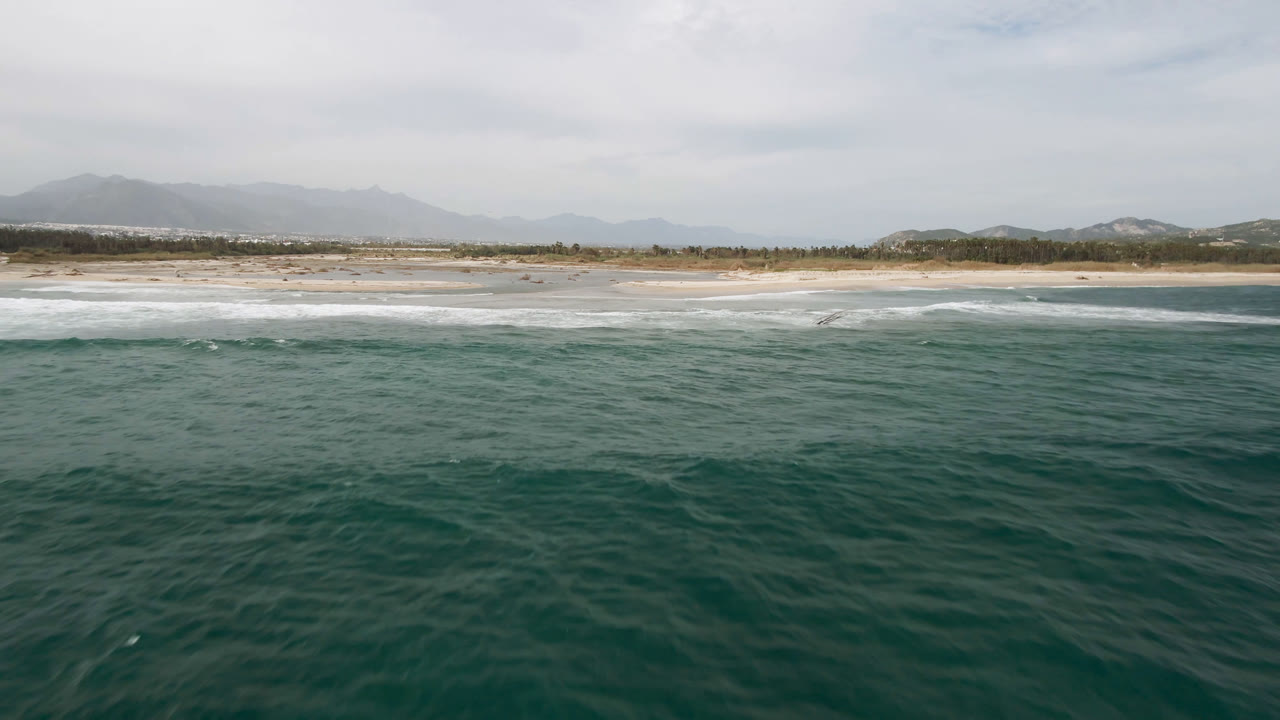 Aerial view of green ocean, white surf, sandy beach, and distant hills under clouds.