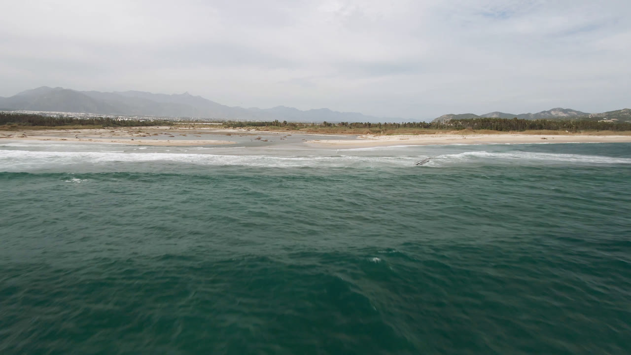 Wide aerial view in San Jose del Cabo with textured green water below, white waves reaching the beach, a thin line of vegetation, and pale mountains in the background.