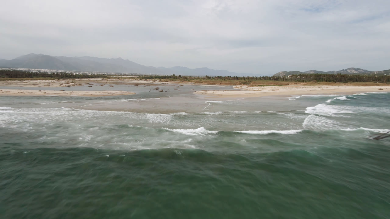 Wide aerial view of green surf meeting a sandy San Jose del Cabo shoreline, with pale sandbars, low coastal vegetation, hazy mountains, cloud cover, and no visible people.