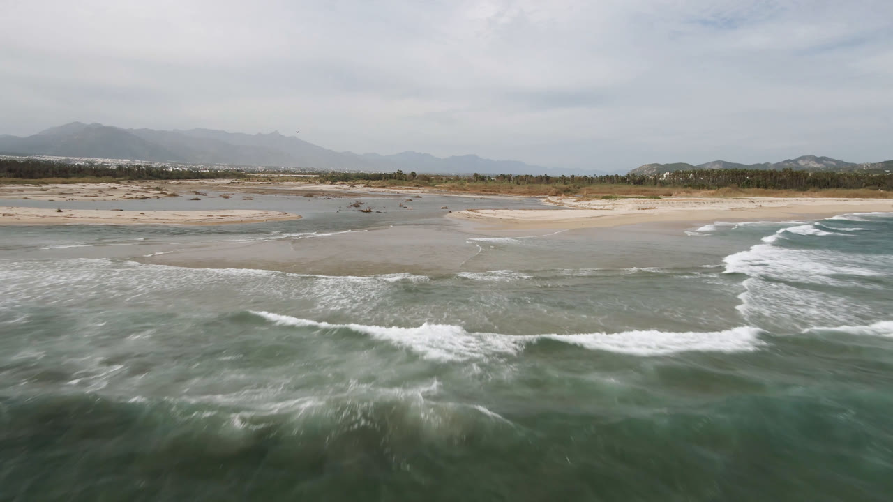 Aerial view over San Jose del Cabo surf and shallow tidal channels, with broad tan sandbars, a low tree line, distant mountains, muted daylight, and no visible people.