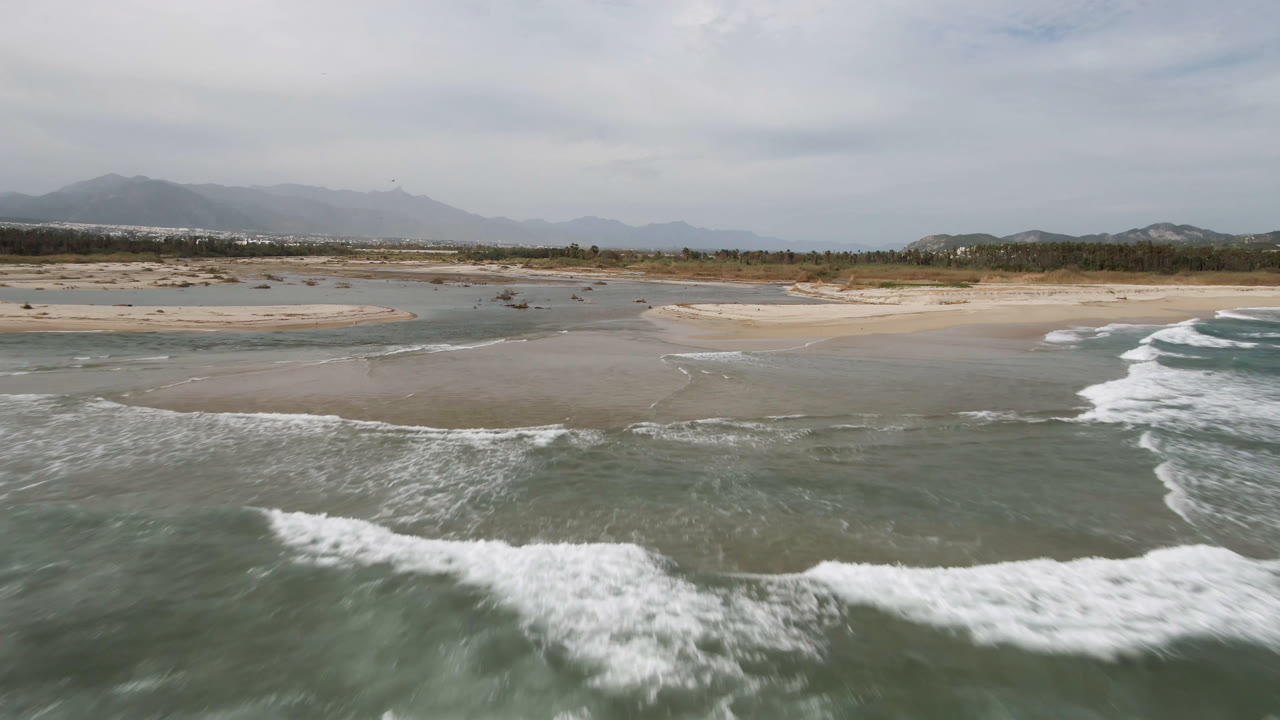 Drone view of waves curling toward a wide beach with sandbars and hazy mountains.