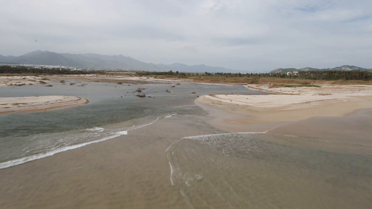 Aerial view of shallow water, pale sandbars, beach surf, and distant mountains.