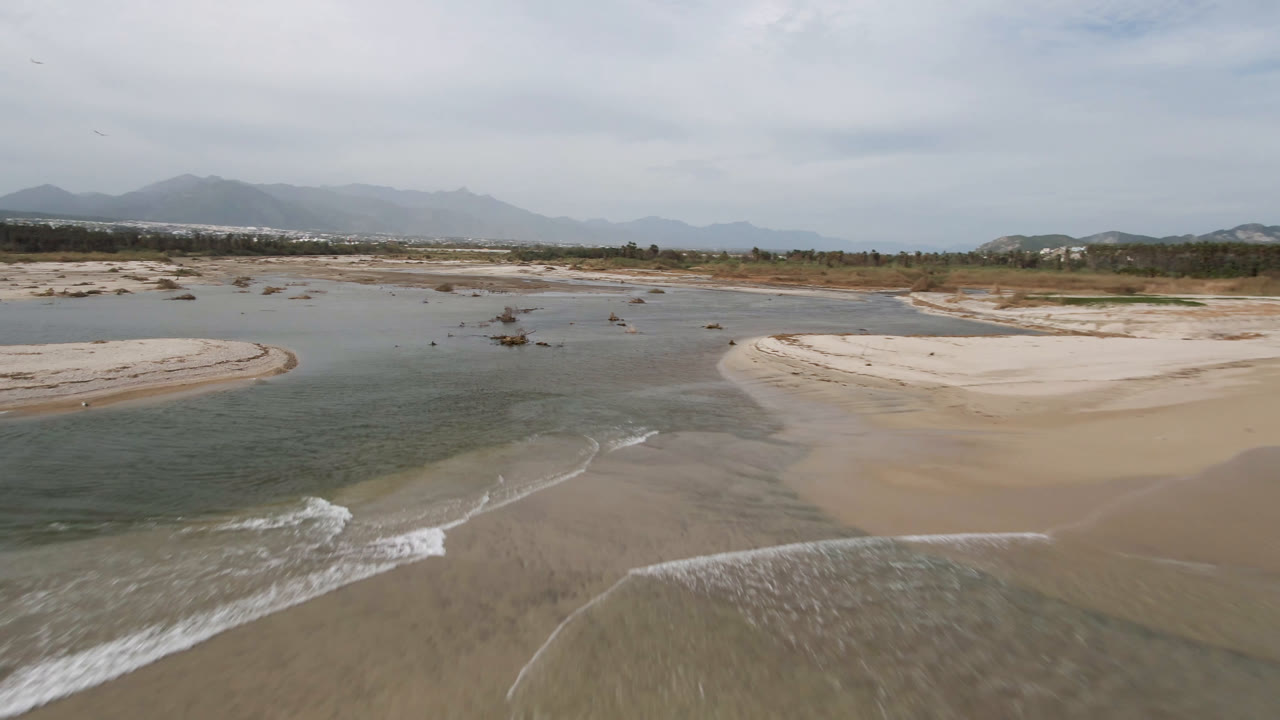 Aerial view in San Jose del Cabo with a curved sandbar centered between green-blue river water and a breaking foreground wave, with no visible people.
