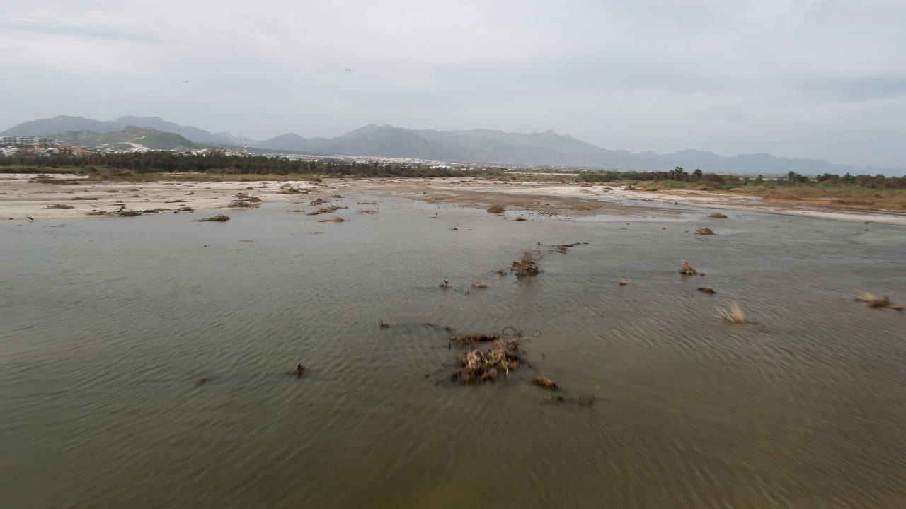 Aerial view over shallow green-gray water and small rock clusters in San Jose del Cabo, Baja California Sur, with hazy mountains on the horizon and no visible people.