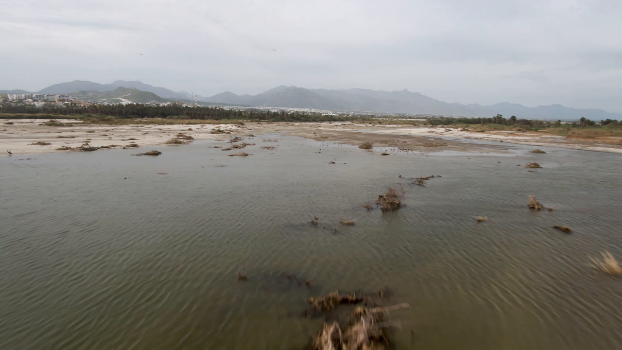 Aerial frame in San Jose del Cabo looking across rippled shallow water, scattered brush-covered rocks, pale flats, distant low buildings, and no visible people.