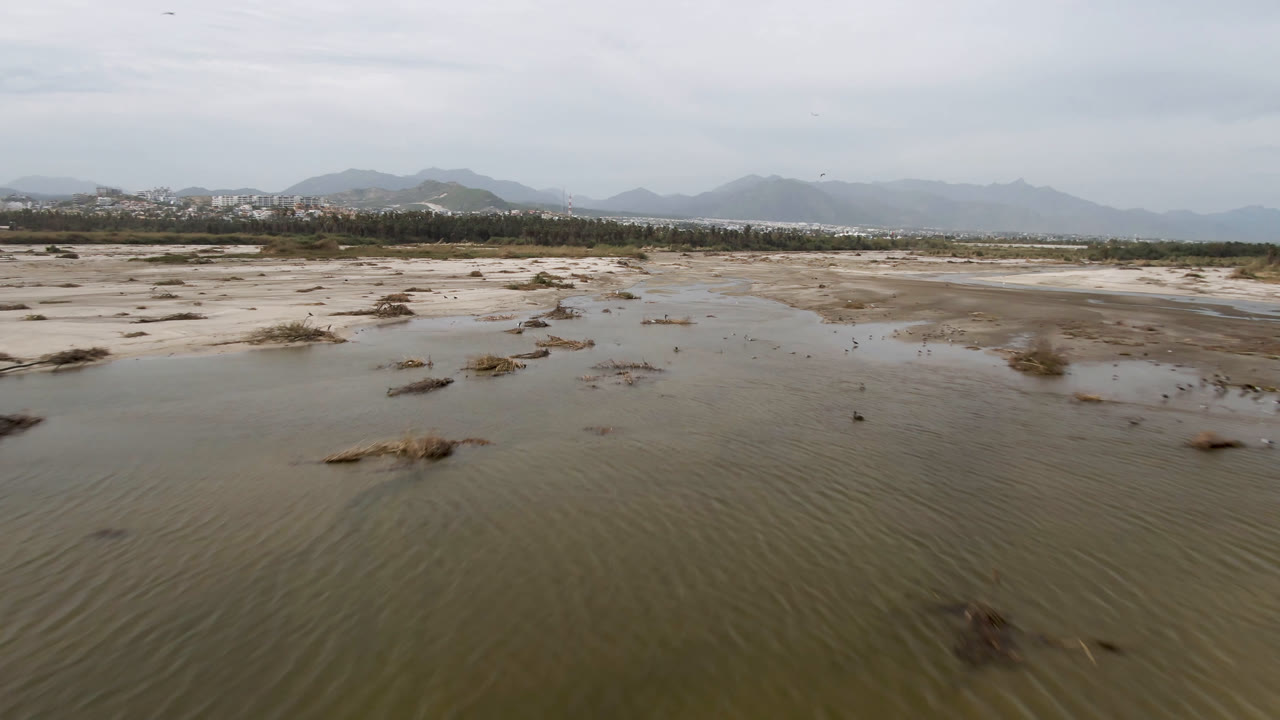 Low aerial view over shallow muddy water and sand flats in San Jose del Cabo, with dry grass clumps in the foreground, mountains on the horizon, and no visible people.