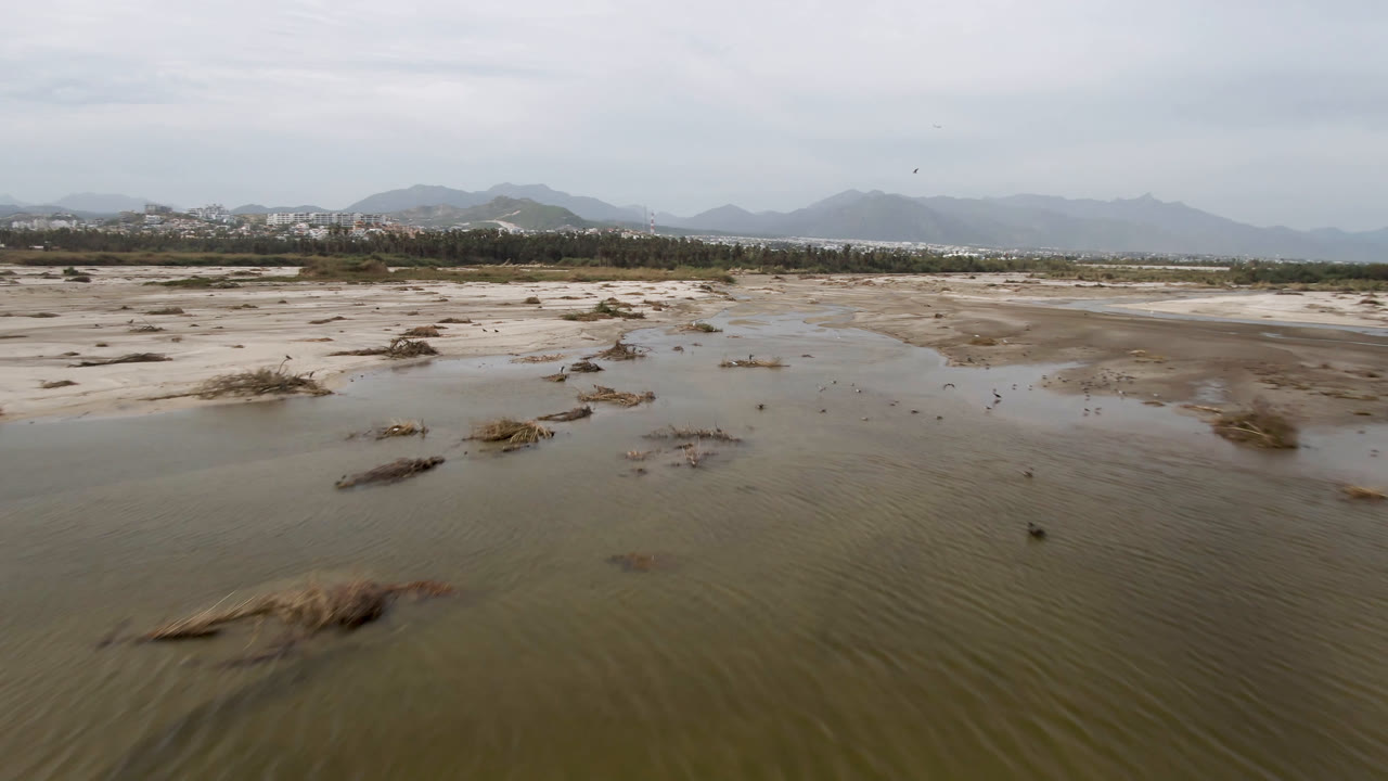 Wide low aerial frame of the San Jose del Cabo muddy beach, with a shallow channel curving through sand, scattered dry grass, hazy mountains, and no visible people.