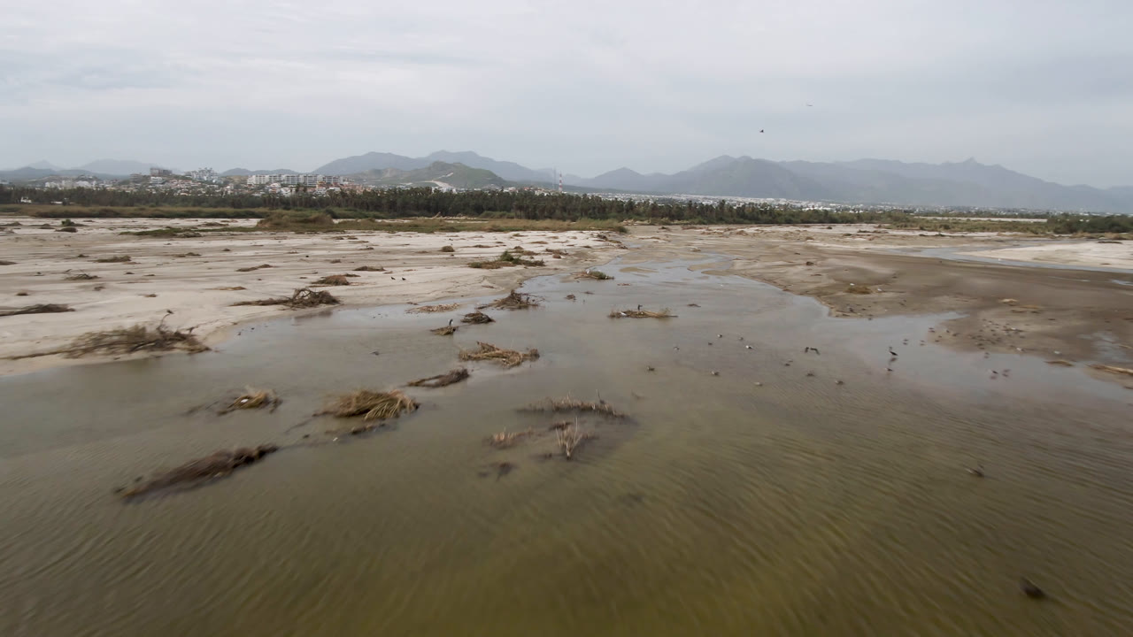 Flat muddy shoreline with shallow water, scattered debris, distant town edge, and mountains.
