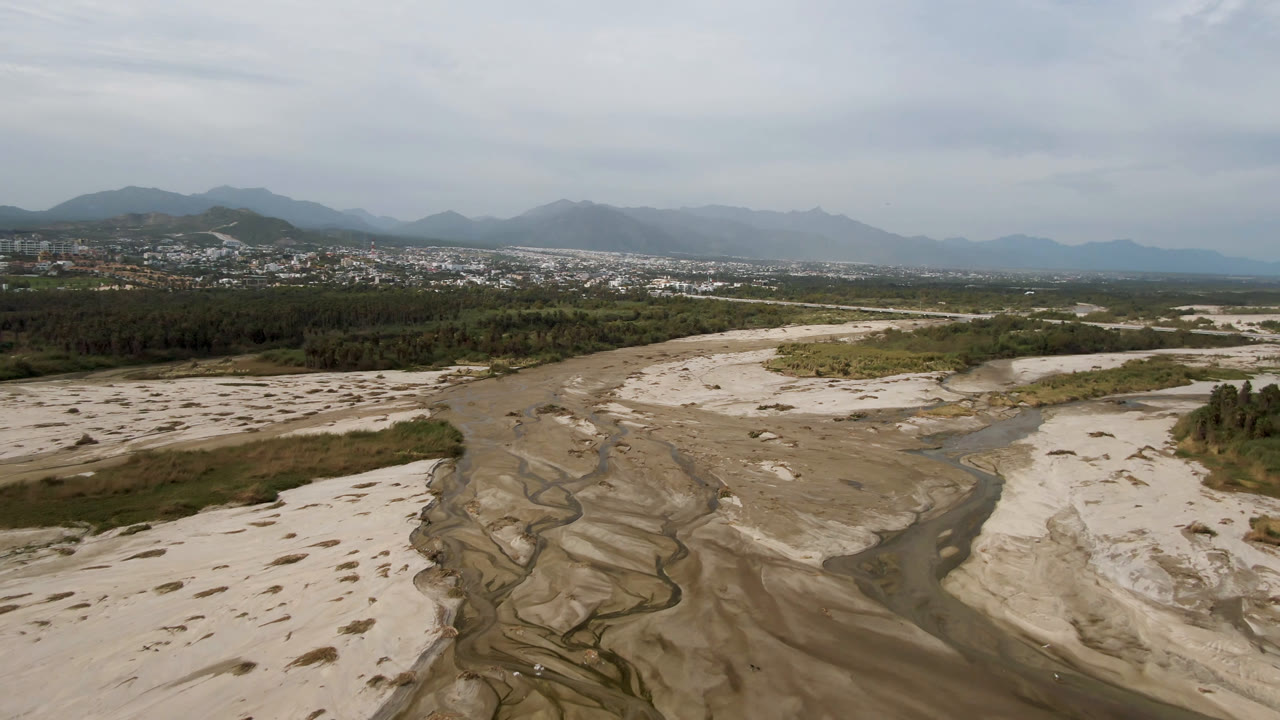 Aerial view in San Jose del Cabo showing pale sand channels, shallow water tracing through the riverbed, a green tree line, distant buildings, mountains, and no visible people.