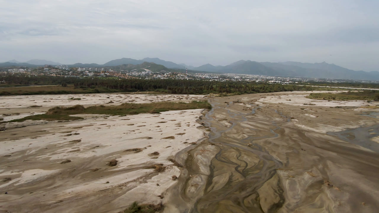 A wide aerial frame in San Jose del Cabo shows branching water paths across the sandy wash, a dark green tree line, distant town buildings, mountains, and no visible people.