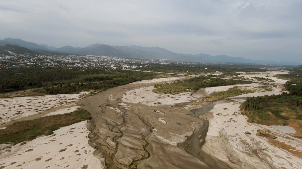 Aerial view of a sandy riverbed, stream channels, vegetation, town, and mountains.