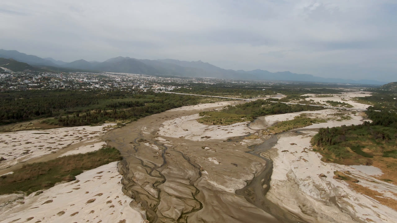 Aerial frame one second in showing the riverbed curving through pale sand toward vegetation and a town at the foot of mountains in San Jose del Cabo, Mexico, with no visible people.