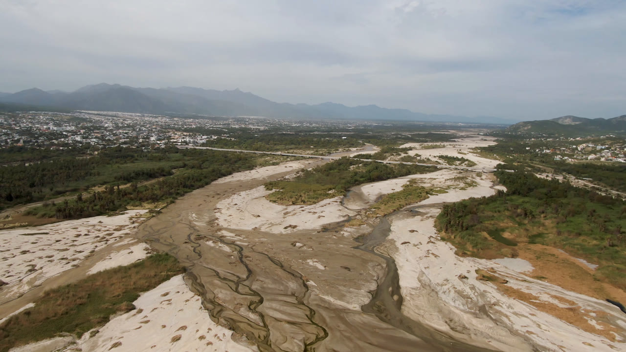 Aerial view of a sandy riverbed winding through a valley toward a distant town and mountains.