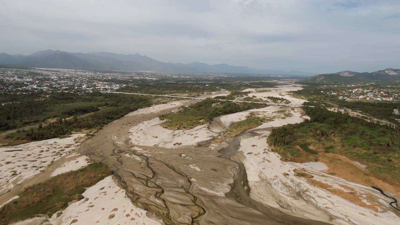 Wide aerial frame in San Jose del Cabo with the sandy riverbed centered, green vegetation on both banks, distant buildings across the valley, hazy mountains, and no visible people.