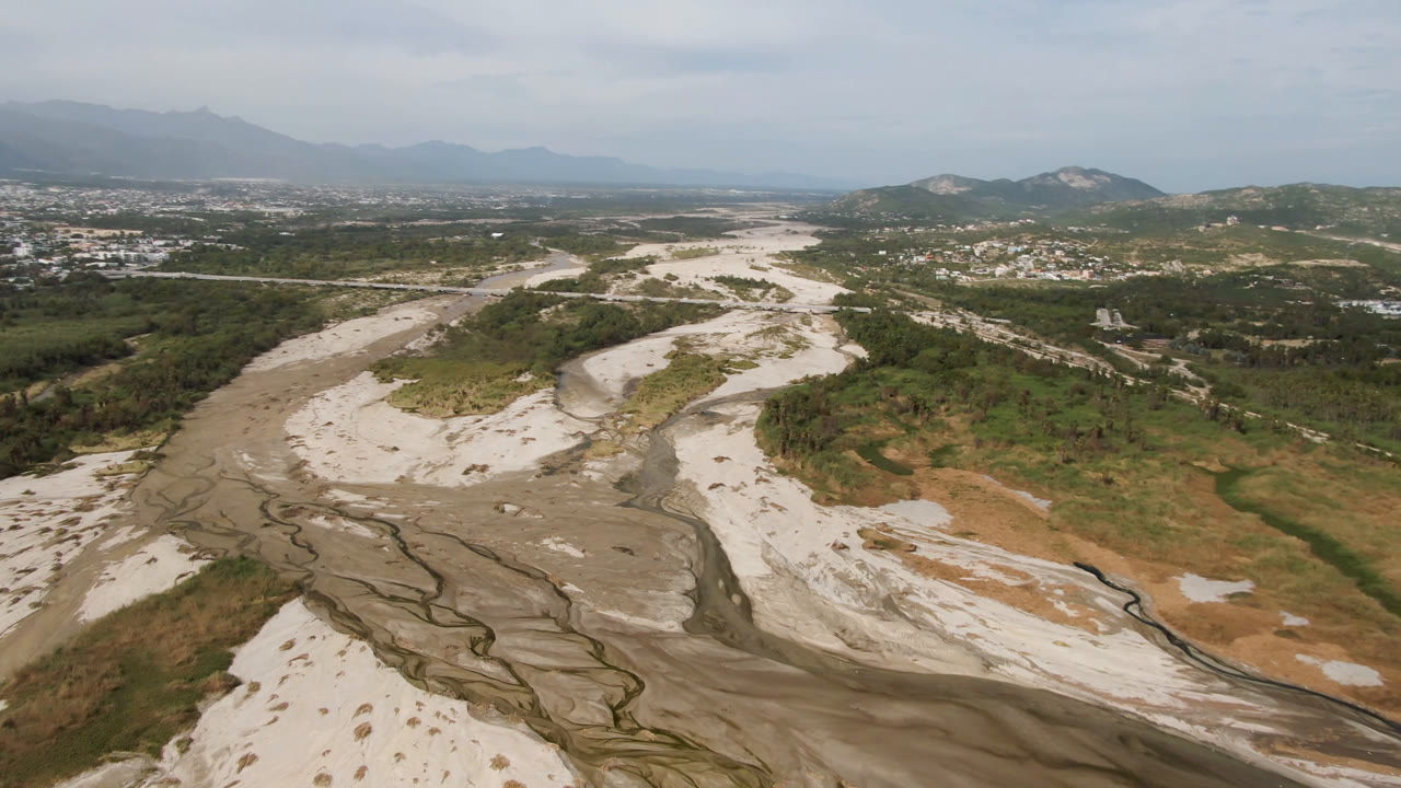 Opening aerial frame over a dry riverbed in San Jose del Cabo, Baja California Sur, with green scrub, pale sand channels, distant hills, town development, and no visible people.