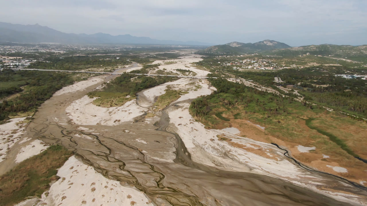 Second aerial frame in San Jose del Cabo showing pale dry channels across scrubland, a road line through the valley, low hills, distant buildings, and no visible people.