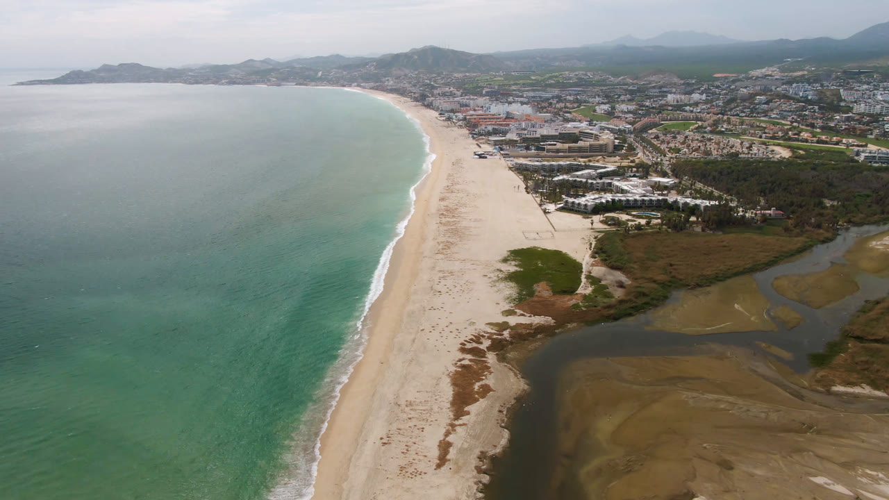 Opening aerial frame over San Jose del Cabo, Mexico, showing turquoise ocean, a long sand beach, lagoon water, coastal buildings, hazy hills, and no visible people.