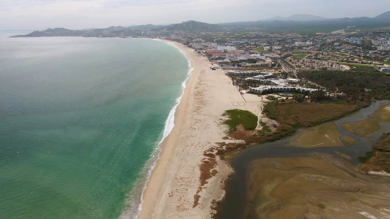 Aerial view in San Jose del Cabo with the shoreline running through the frame, ocean to the left, lagoon and wetland textures to the right, distant buildings, and no visible people.