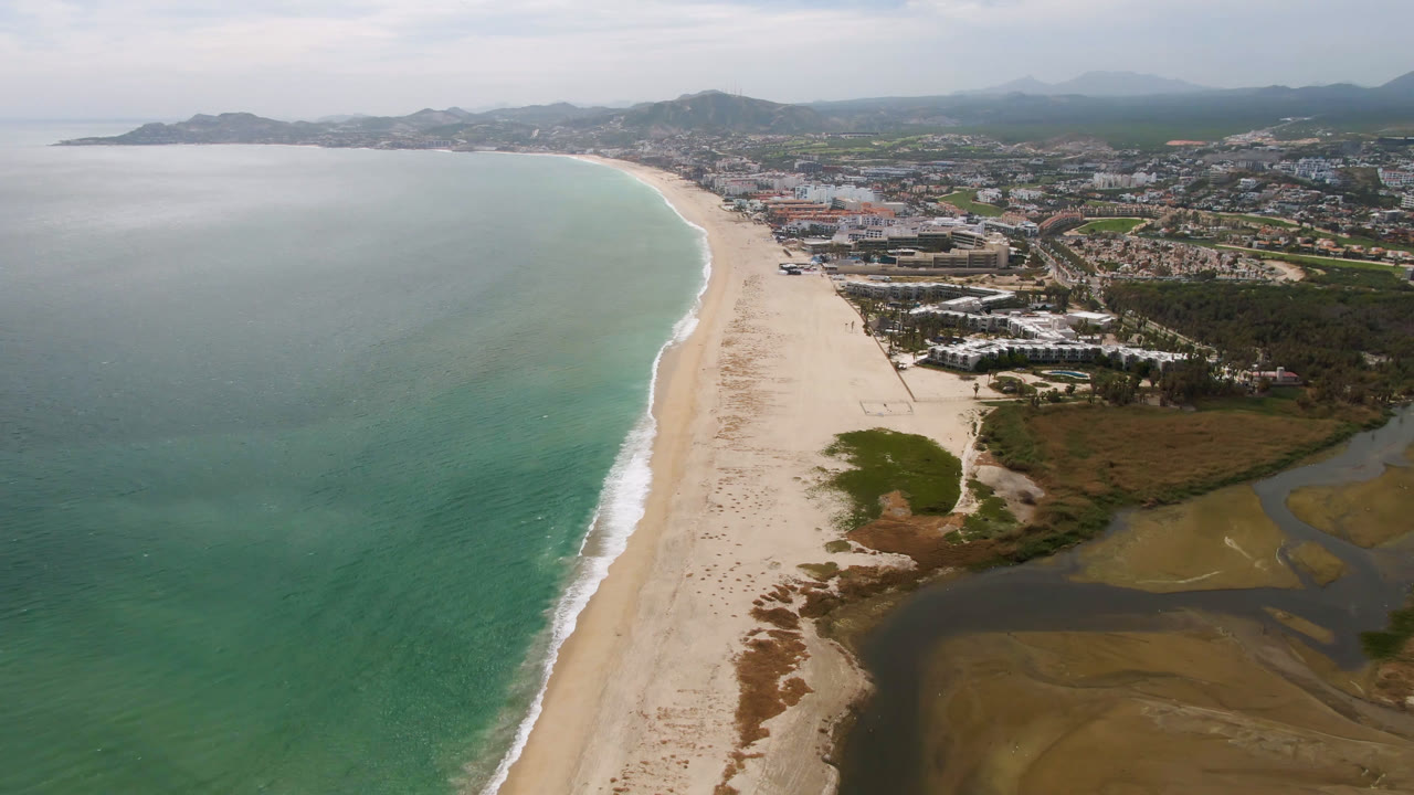 Aerial view over San Jose del Cabo with turquoise ocean at left, a long sand beach through the center, lagoon water at lower right, inland buildings, distant mountains, and no visible people.