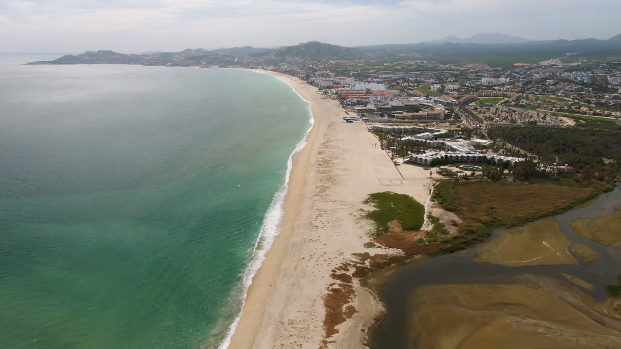 Drone-height view in San Jose del Cabo showing the curved surf line along the beach, green-blue ocean to the left, wetland water at bottom right, low urban buildings inland, and no visible people.