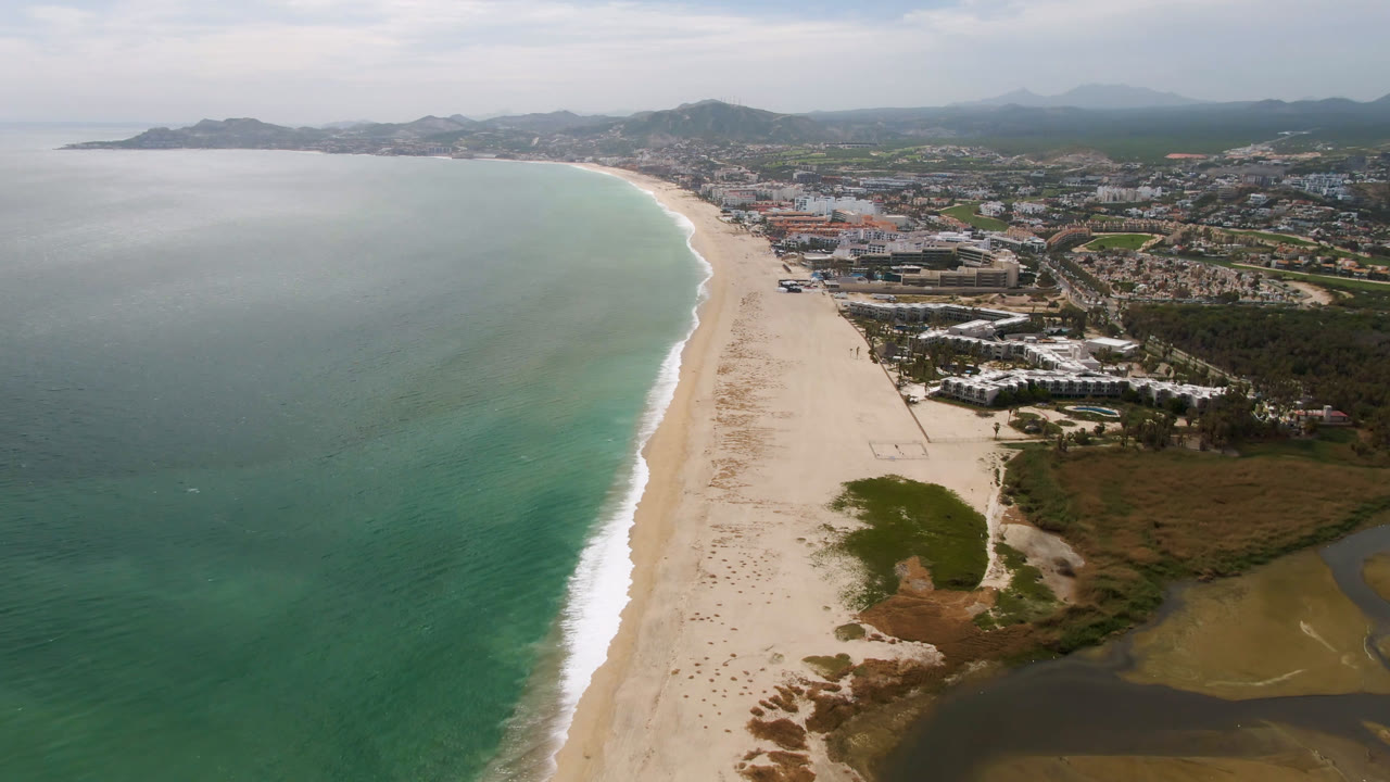 High aerial view over San Jose del Cabo with teal ocean on the left, a long sand beach through the center, inland buildings to the right, distant hills, and no visible people.