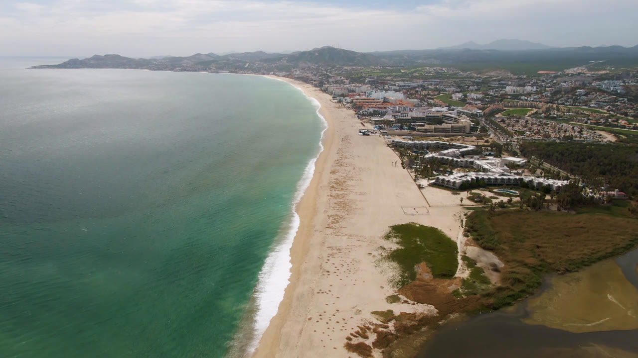 Aerial frame from San Jose del Cabo showing the beach running diagonally beside green-blue water, with low coastal buildings, roads, and hazy mountains beyond, and no visible people.