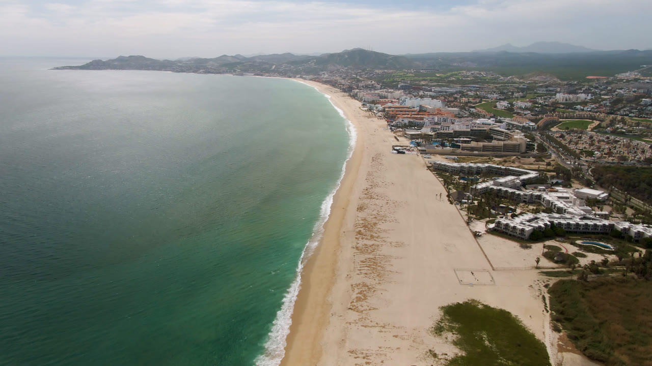 Opening aerial frame over San Jose Del Cabo, Mexico, with green-blue ocean on the left, a pale curving beach through the center, coastal buildings to the right, distant hills, and no visible people.