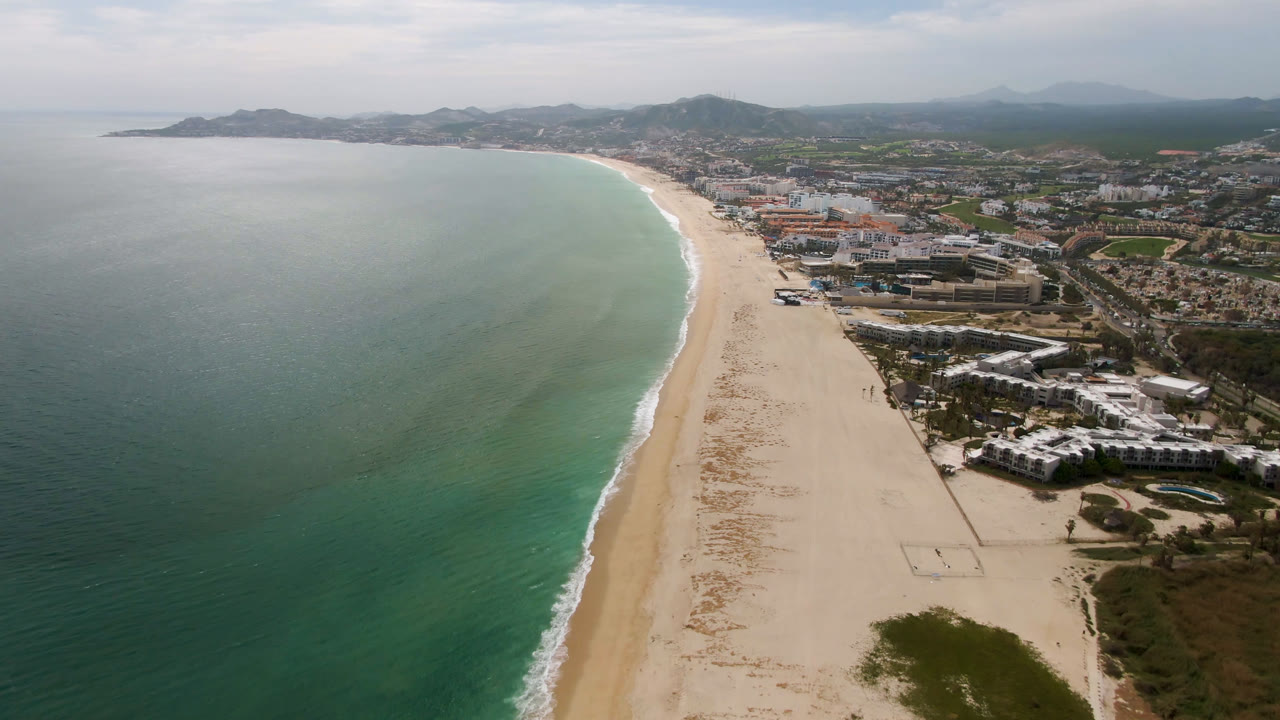 Second aerial frame from San Jose Del Cabo, Mexico, showing more of the long sandy beach, the ocean edge, clustered shoreline buildings, inland roads, hazy hills, and no visible people.