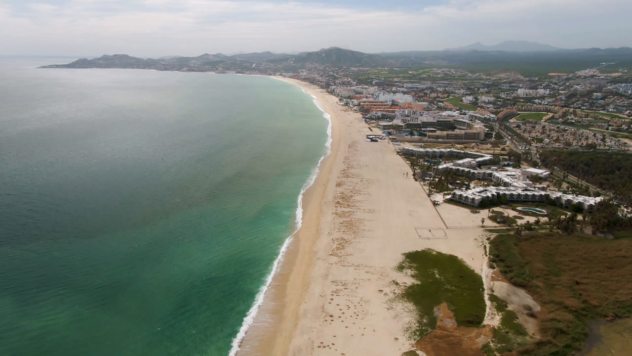 Aerial view of turquoise ocean beside a long sand beach and coastal buildings.