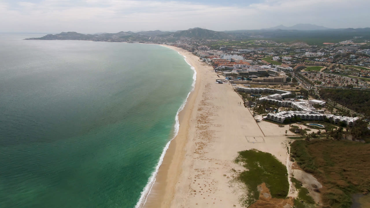 Aerial frame over San Jose del Cabo with green-blue ocean filling the left side, a narrow surf edge, pale sand running into the distance, clustered buildings on the right, and no visible people.