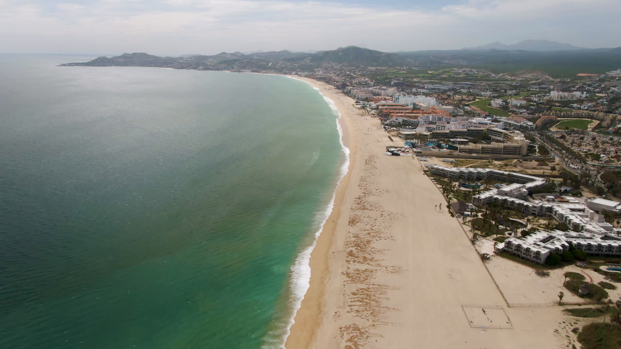 Opening aerial frame over San Jose del Cabo, Mexico with turquoise water beside a long pale beach, coastal buildings to the right, hazy hills beyond, and no visible people.