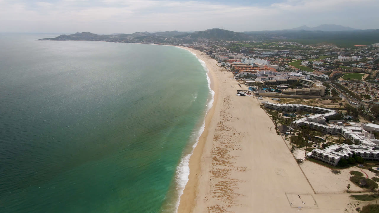 Second frame of San Jose del Cabo coastline showing more of the pale beach curve, green-blue ocean water, clustered coastal buildings, roads, distant hills, and no visible people.