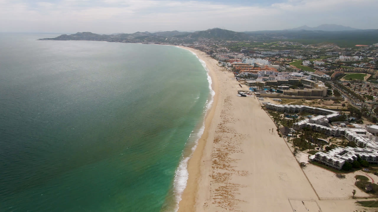 Drone view down a long beach with ocean on the left and resort-like buildings on the right.
