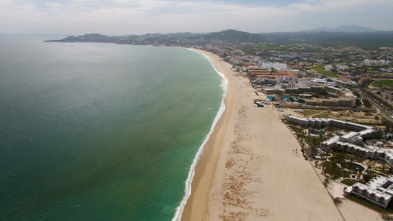 Aerial frame from San Jose del Cabo looking along the curved shoreline, with darker ocean water at left, a narrow surf edge, city blocks at right, hazy hills, and no visible people.
