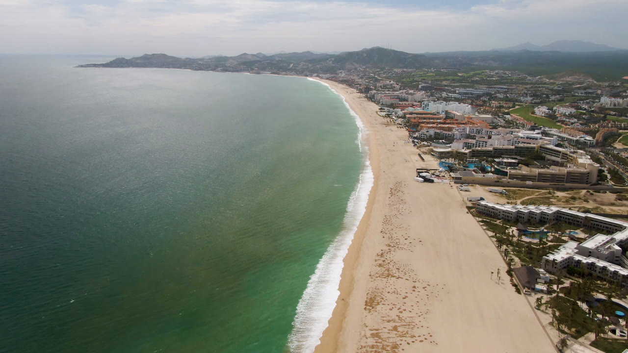 Aerial view over San Jose Del Cabo with green-blue ocean beside a wide sandy beach, coastal buildings on the right, hazy hills in the distance, and no visible people.