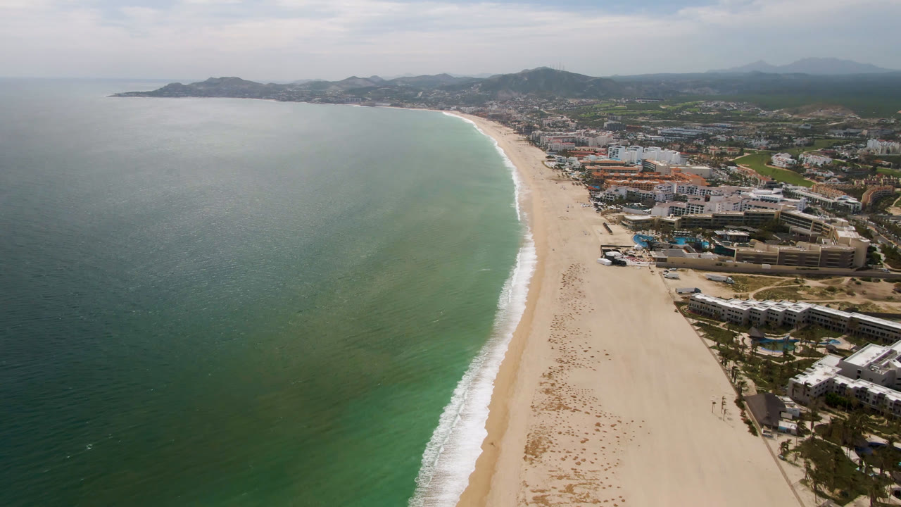 High aerial frame from San Jose Del Cabo showing the beach curving along green-blue water, with low buildings tight to the shore and no visible people.
