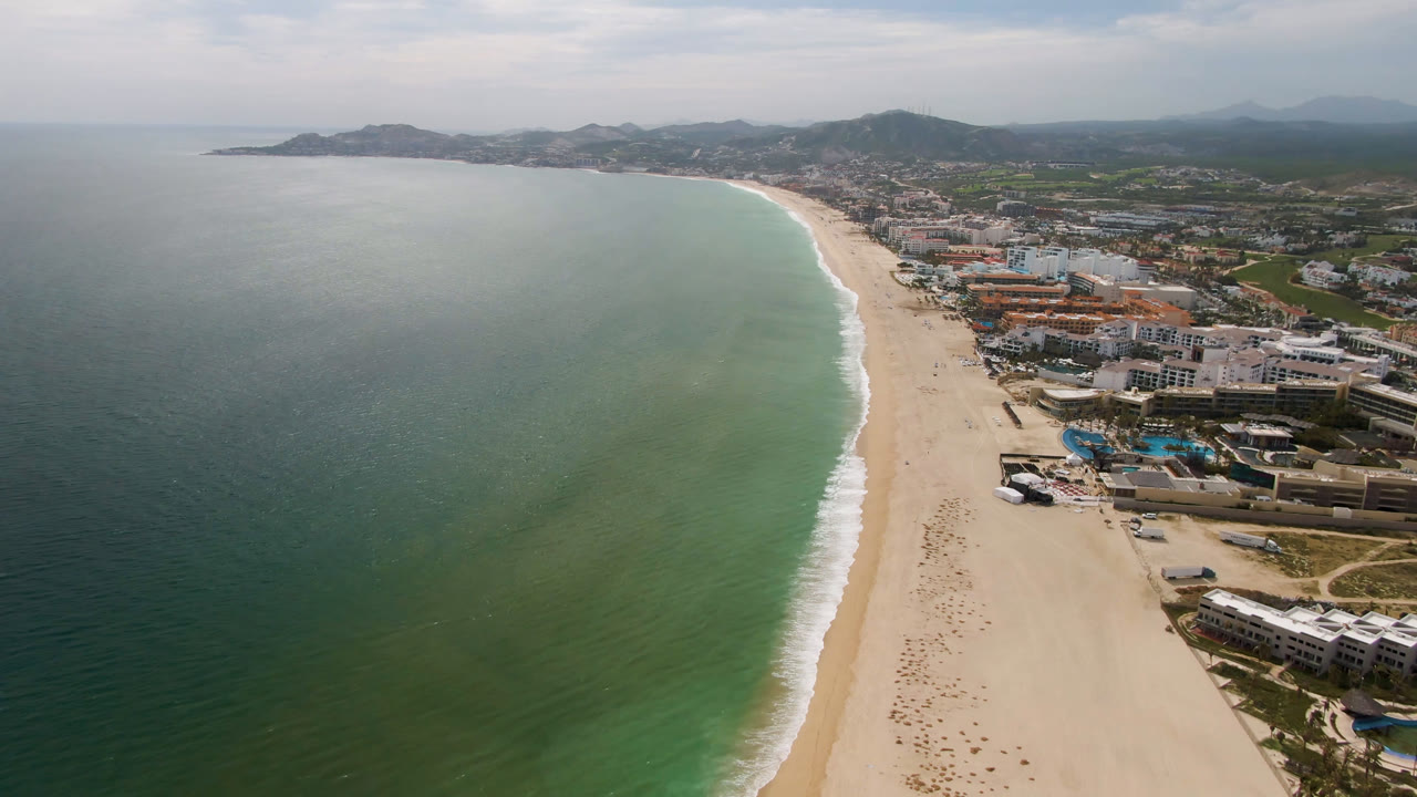 High aerial view over San Jose del Cabo, Mexico, with green ocean on the left, a pale curving beach in the center, coastal buildings on the right, distant hills, and no visible people.